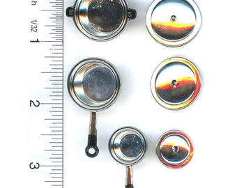 May include: A set of three miniature metal cooking pots and pans with matching lids, arranged next to a vertical ruler showing measurements in inches. The cookware features silver-toned metallic surfaces and black handles on the two pans.