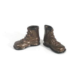 May include: A pair of miniature brown boots with laces, set against a white background. The boots have a metallic finish and are detailed with stitching and eyelets. The boots are small, likely for a doll or decorative purpose.