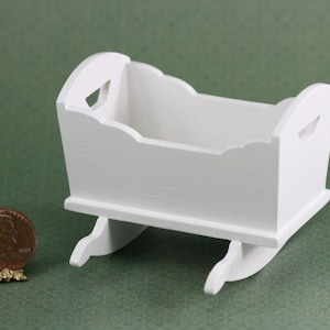 May include: A small, white wooden cradle with a scalloped top edge and curved rockers. The cradle has cut-out handles on each end. A penny is next to the cradle for scale.