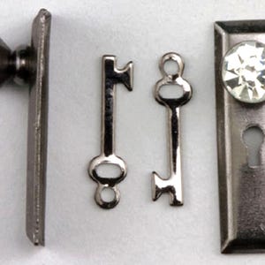 May include: A set of miniature dollhouse hardware, including a doorknob, a keyhole plate with a crystal knob, and two small keys. The hardware has a dark, metallic finish, and the crystal knobs sparkle. The items are designed for a dollhouse or miniature project.