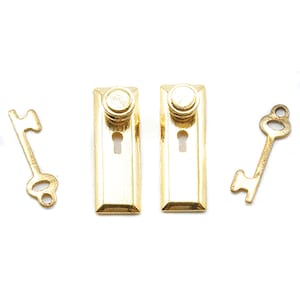 May include: A set of gold-colored miniature door hardware, including two rectangular door plates with keyholes and two decorative keys. The hardware has a shiny, reflective surface, suggesting a vintage or antique style.