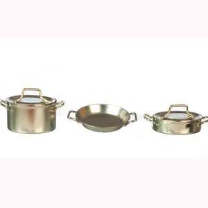 May include: A set of three miniature metal cooking pots and pans. The set includes a lidded pot with a gold handle, a small frying pan, and a second lidded pot with a gold handle. The pots and pans are silver in color.