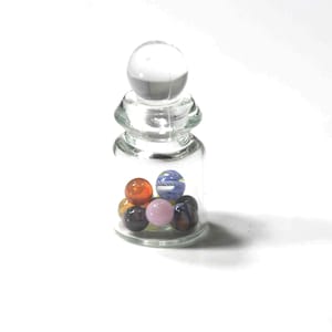 May include: A clear glass bottle with a glass stopper, filled with colorful marbles. The marbles are various colors, including orange, blue, pink, and black. The bottle is on a white background.