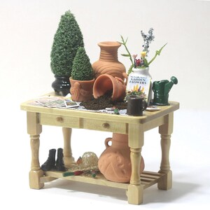 May include: Miniature garden scene on a light wood table. Includes terracotta pots, a small watering can, a vase with flowers, and a small black boot. The table has a lower shelf with a large terracotta pot and other gardening accessories.