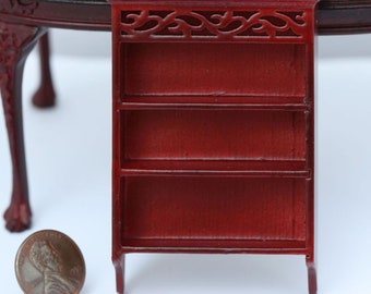 May include: Miniature red bookshelf with three shelves and decorative top. The small bookcase is made of wood and has a classic design. A penny is next to the bookshelf for size comparison. The bookshelf is part of a dollhouse furniture set.