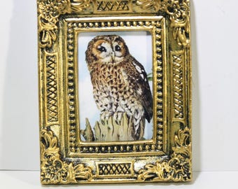 May include: A small, rectangular, gold-colored picture frame with an ornate, embossed design. The frame surrounds a print of a brown and white owl perched on a tree stump. The frame has a detailed, textured surface.