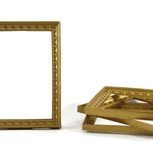 May include: A collection of small, ornate, gold-colored picture frames. One frame stands upright, while several others are stacked. A penny is included for scale, showing the frames' miniature size. The frames have decorative edges.