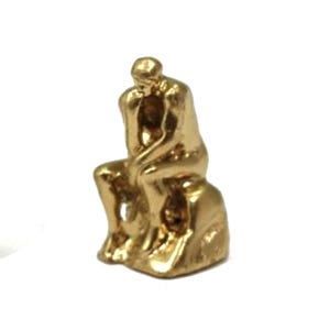 May include: A small, gold-colored sculpture of a seated figure in a contemplative pose. The figure is resting their chin on one hand, with the other hand on their leg. The sculpture is on a small, textured base.