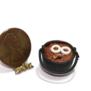 May include: Miniature black cauldron filled with a brown stew, topped with white rings. A copper penny is next to the cauldron for scale, showing the small size of the food item. The cauldron has two black handles.