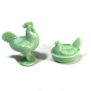 May include: Two light green chicken figurines. One is a rooster standing on a pedestal base. The other is a hen sitting in a nest. Both are made of a solid, smooth material.
