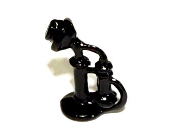 May include: A small, glossy black figurine shaped like a vintage candlestick telephone. The object features a round base, a central vertical column, a curved mouthpiece at the top, and a separate receiver resting on a side hook.
