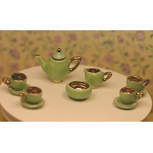 May include: A miniature tea set in a light green color with gold trim. The set includes a teapot, creamer, sugar bowl, and several teacups with saucers. The tea set is arranged on a white surface.