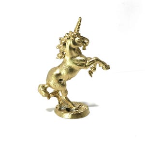 May include: A gold-colored unicorn figurine rearing up on its hind legs. The mythical creature has a textured surface, a spiraled horn, and a flowing mane. The unicorn is mounted on a small, textured base.
