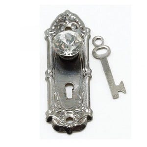 May include: A silver-toned miniature door knob set with a clear, faceted crystal knob and a matching key. The door plate has ornate detailing. The key is a simple, skeleton key design.