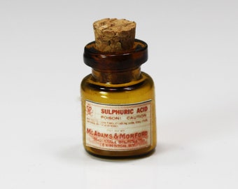 Puede incluir: Pequeña botella de vidrio ámbar con tapón de corcho. La etiqueta dice "SULPHURIC ACID POISON! CAUTION" y "McADAMS & MORFORD".