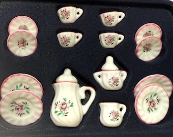 May include: A miniature porcelain tea set with a floral rose design. The set includes cups, saucers, a creamer, a sugar bowl, and a teapot. Each piece is white with pink roses and green leaves, and pink trim.