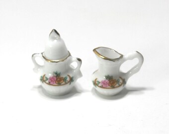 May include: Miniature white porcelain creamer and lidded sugar bowl set. Both pieces have gold trim and a floral design with pink and orange roses. The sugar bowl has two handles and a pointed lid. The creamer has a handle and a spout.