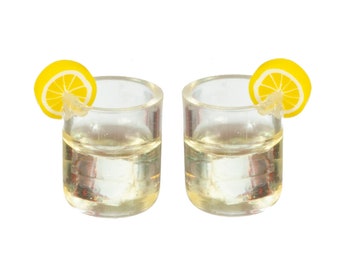 May include: Two clear shot glasses filled with a light-coloured liquid, each garnished with a yellow lemon slice. The glasses are cylindrical and appear to be made of plastic or glass. The background is plain white.