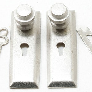 May include: Two silver-colored door handle plates with keyholes and a knob on top. Two matching silver keys are placed on either side of the door handle plates. The items are likely for home decor.