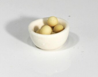 May include: A small, off-white ceramic bowl filled with several light yellow, oval-shaped objects. The bowl has a rounded shape and sits on a white surface. The objects inside appear to be small, possibly food items.