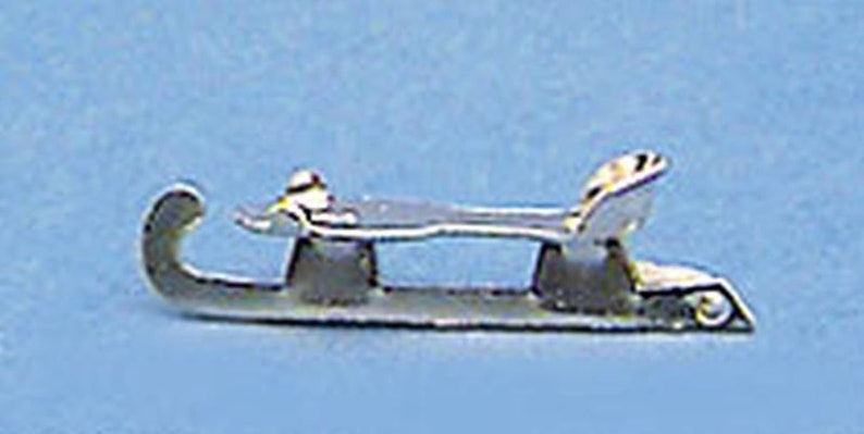 May include: A miniature silver-colored sled, likely a charm or pendant. The sled has curved runners, a flat seat, and a small backrest. The design is simple and elegant, with a focus on the sled's form.
