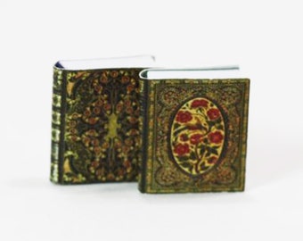 May include: Two miniature books with ornate, antique-style covers. One book features a floral design in gold, green, and red. The other book has a similar design with a central oval of red flowers and a bird. The books are set against a white background.