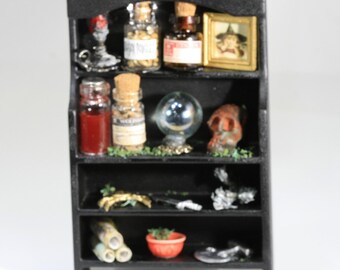 May include: A miniature black shelf unit filled with various items. It features small glass bottles with corks, a crystal ball, a framed picture, a skull, and other decorative elements. The shelf is designed to resemble a witch's cabinet.