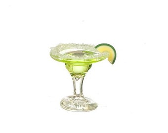 May include: A margarita cocktail in a clear glass with a green liquid, a salt-rimmed edge, and a lime wedge. The glass has a wide bowl and a stem. The drink is set against a white background.