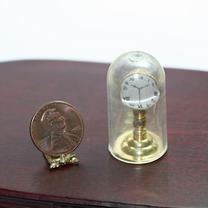 May include: A miniature clock with a silver face and Roman numerals, encased in a clear glass dome, next to a penny coin. The clock has a gold-colored base. The penny is displayed on a small gold-colored stand.