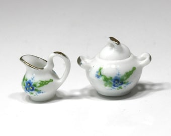 May include: Miniature white porcelain creamer and sugar bowl set. The set features a gold trim and is decorated with blue floral designs and green leaves. The creamer has a handle and spout, while the sugar bowl has a lid.