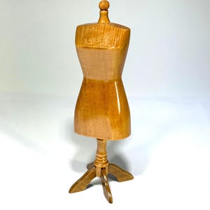 May include: A miniature dress form mannequin crafted from polished light brown wood. The torso features a smooth, contoured shape with a small knob at the neck, supported by a central post and a three-legged base stand for stability.