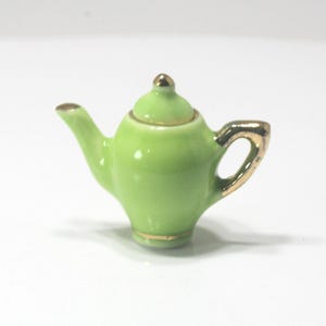 May include: A small, lime green ceramic teapot with gold trim. The handle, spout, and lid are accented with gold. The teapot has a glossy finish and a small, rounded lid with a gold knob. A miniature decorative item.
