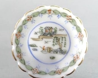 May include: A small, white porcelain dish with a scalloped edge and gold trim. The center features a landscape scene with a house, trees, and figures, framed by a blue border. The rim is decorated with green leaves and orange floral accents.
