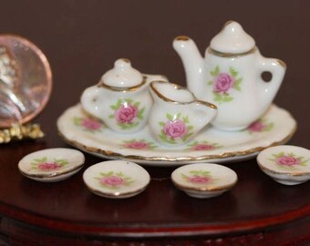 May include: Miniature white porcelain tea set with pink rose details, including a teapot, creamer, sugar bowl, and saucers. The set is displayed on a decorative plate with gold trim. A penny is next to the tea set.
