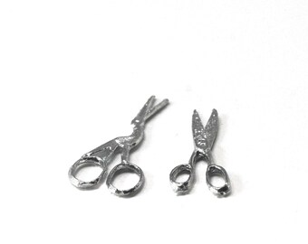May include: Two small, silver-colored metal charms shaped like scissors. One pair of scissors is positioned at an angle with open blades, while the other pair is positioned vertically with closed blades. Both items feature circular finger loops.