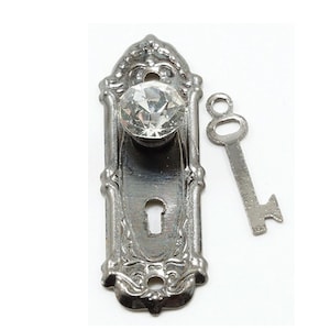 May include: An antique-style door handle set with a clear crystal knob and a matching key. The handle plate is silver-toned with ornate detailing. The key is a simple, vintage design. The set is likely for a small cabinet or decorative use.