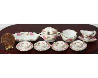 May include: Miniature porcelain tea set with pink floral designs. The set includes plates, bowls, a serving dish, a gravy boat, and a lidded soup tureen. The set is displayed on a dark wooden surface.