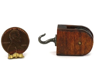 May include: A miniature wooden pulley block with a dark brown finish and a silver-toned metal hook, positioned next to a 2014 United States copper penny for scale, showing the small size of the mechanical component.