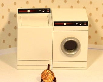 May include: Miniature white washing machine and dryer set with black and red accents. The washer has a top-loading design, while the dryer features a front-loading door. A penny is in the foreground, providing a sense of scale. The set is on a white surface.