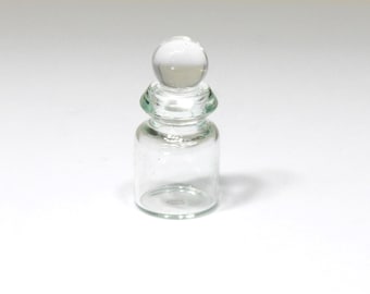 May include: A small, clear glass bottle with a rounded base and a spherical stopper. The bottle has a short neck and a slightly wider body. The stopper is a clear, round ball shape. The bottle is empty and sits on a white surface.
