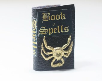 May include: Miniature black book with the words "Book of Spells" in gold lettering. A gold spider charm is affixed to the front cover. The spine has gold lettering. The book is a prop or decorative item.