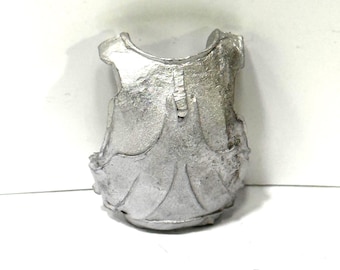 May include: A small, silver-colored metal chest plate with a textured surface. The miniature armor piece has a curved neckline and a vertical ridge down the center, with decorative details along the bottom edge.