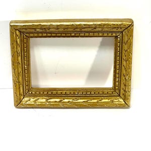 May include: A rectangular gold-colored picture frame with a detailed, textured surface. The frame has a rectangular opening in the center and is likely made of wood. The frame's design includes small, repeating patterns along the edges.