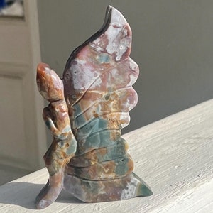 May include: A carved stone sculpture of a butterfly with wings in shades of pink, brown, green, and white. The sculpture is made of agate and has a natural, organic look.
