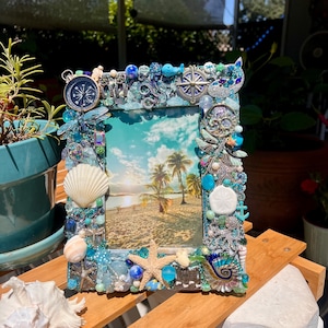 May include: A rectangular picture frame decorated with an assortment of blue, green, and clear beads, seashells, a starfish, a compass, and a whale figurine. The frame surrounds a photograph of a tropical beach scene with palm trees at sunset.