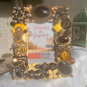 Puede incluir: Un marco de fotos amarillo decorado con joyas negras y doradas, pedrería y una abeja. El marco mide 8,89 cm por 12,70 cm y tiene el texto "Embrace the Journey" en su interior.