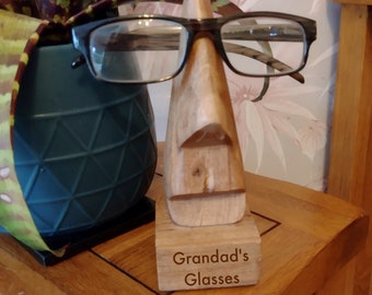 May include: Wooden stand with a carved face holding a pair of eyeglasses. The stand has a wooden base with the text "Granddad's Glasses" engraved on it.