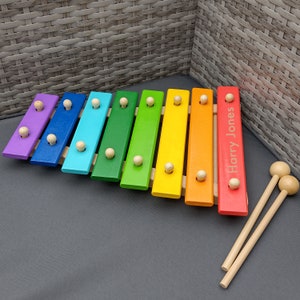 Op de afbeelding: Een regenboogkleurig houten xylofoon met de gepersonaliseerde naam "Harry Jones" op de rode balk. De xylofoon wordt geleverd met twee houten hamers.