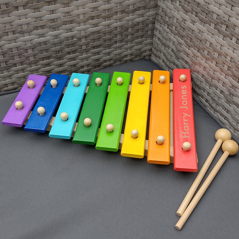 Op de afbeelding: Een regenboogkleurig houten xylofoon met de gepersonaliseerde naam "Harry Jones" op de rode balk. De xylofoon wordt geleverd met twee houten hamers.