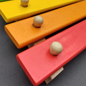 Op de afbeelding: Een houten xylofoon met gekleurde latten en ronde houten hamers. De latten zijn geverfd in tinten geel, oranje en rood.
